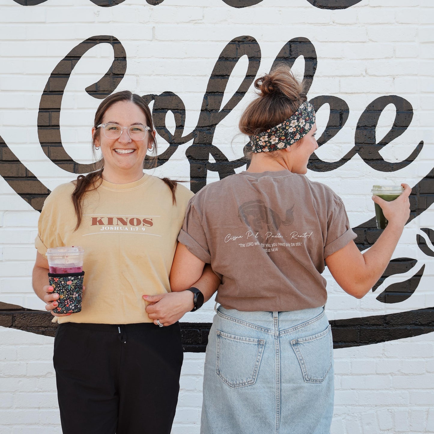 Two women standing in front of a 'Kinos Coffee' mural, holding coffee cups.