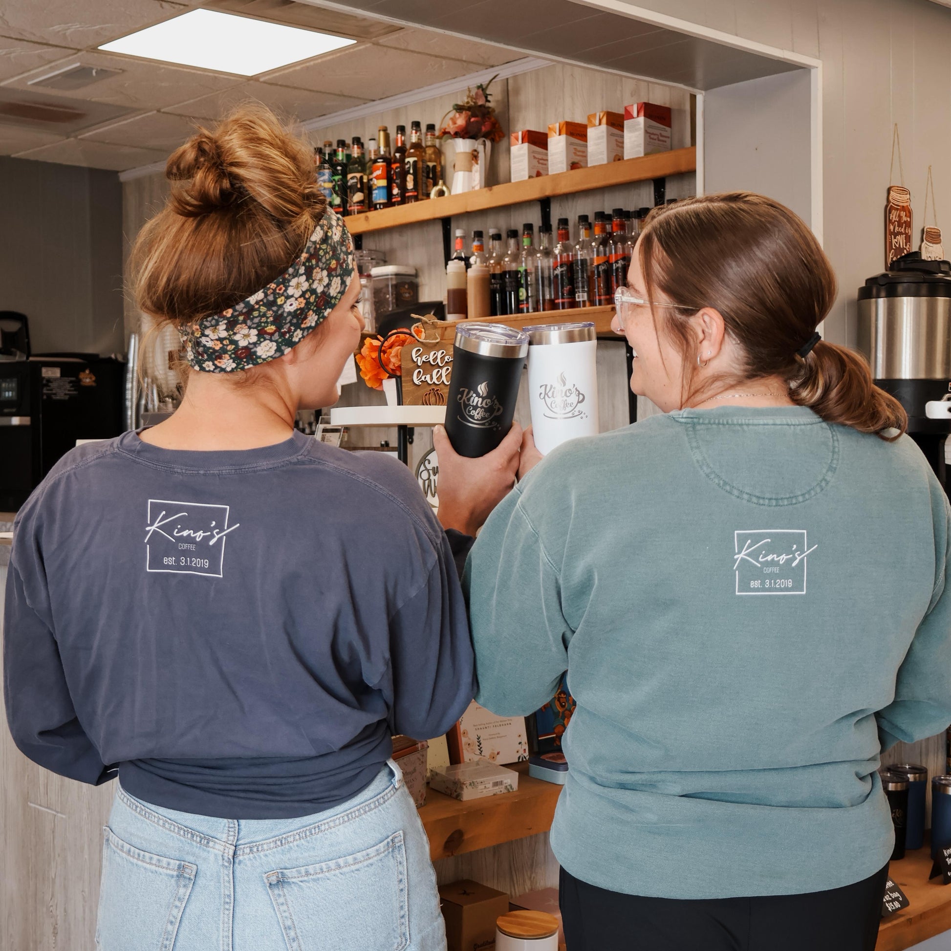 Two women in a kitchen or bar setting, one holding a black cup.